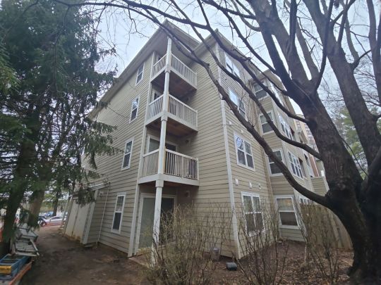 A multi-storey residential apartment building with white and grey siding and private balconies, photographed from close range beneath mature trees on an overcast day.