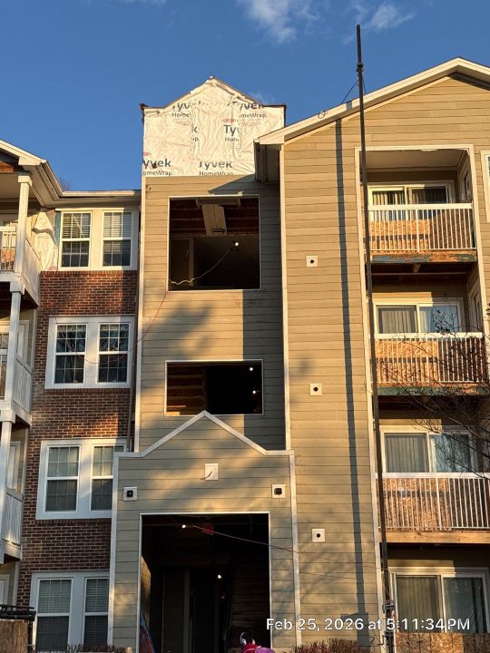 A multi-storey residential apartment building with brick exterior and private balconies with metal railings, photographed from below at a low angle in warm sunlight.
