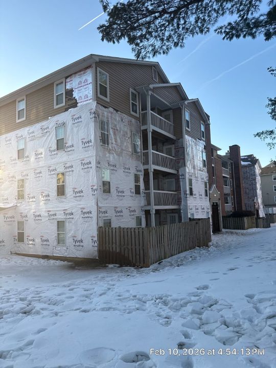 A multi-storey residential apartment building with light grey siding and open balconies, photographed from the exterior in winter with snow covering the ground and surrounding trees.