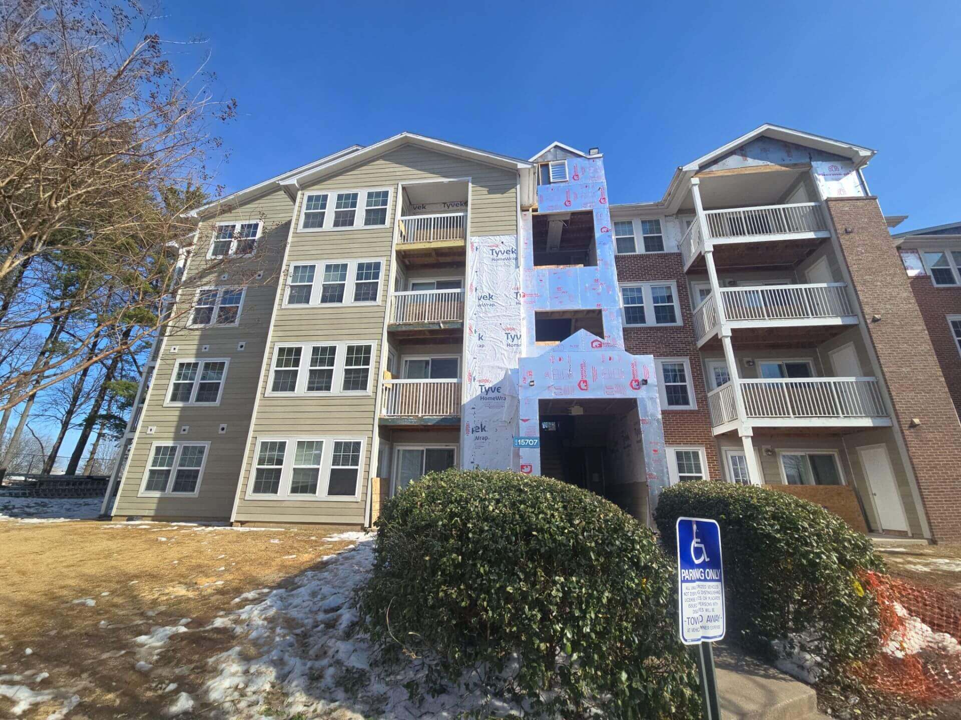 A multi-storey residential apartment building with white and grey siding, featuring private balconies and a covered main entrance, surrounded by landscaped shrubs and photographed on a bright sunny day.