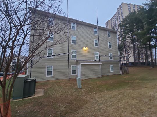 A multi-storey residential apartment building with grey siding, photographed from the side elevation at dusk, with a grassy lawn, bare trees, and a dumpster enclosure visible in the foreground.