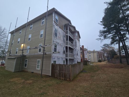 A multi-storey residential apartment building with grey siding and private balconies, photographed from the side along a dirt path, flanked by tall pine trees on an overcast day.