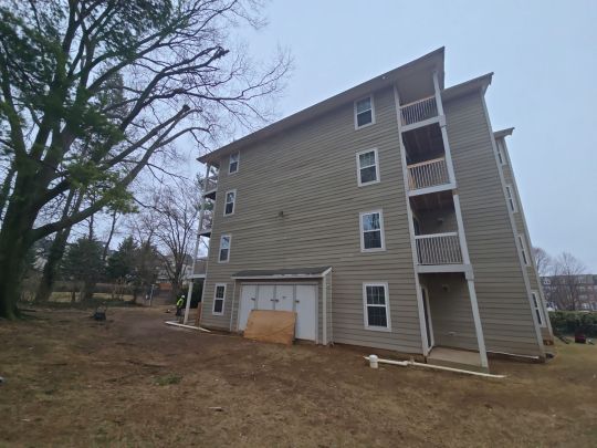 A multi-storey residential apartment building with tan siding and private balconies, photographed from the rear corner across a sloped grassy lawn, with bare trees visible on an overcast day.