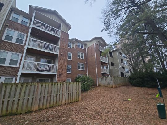 A multi-storey residential apartment building with brick exterior and private balconies, photographed from the rear corner surrounded by mature trees with autumn foliage on the ground and a wooden fence in the foreground.