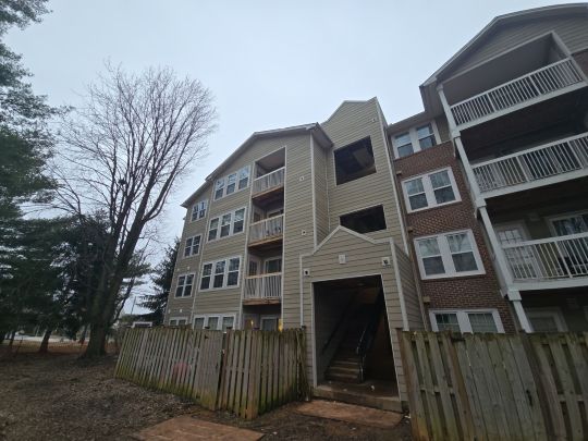 A multi-storey residential apartment building with brick and grey siding, photographed from behind a wooden fence, with bare trees visible against an overcast sky.