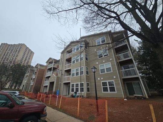 A multi-storey residential apartment building with brick and tan siding, featuring private balconies and a low brick perimeter wall, photographed from the street with bare trees and parked cars visible on an overcast day.